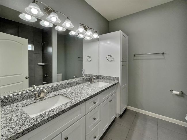 The primary bathroom features a sleek design with a double sink vanity topped with luxurious granite, modern lighting, and a bathtub/shower combo with elegant tile surround. | Image 10