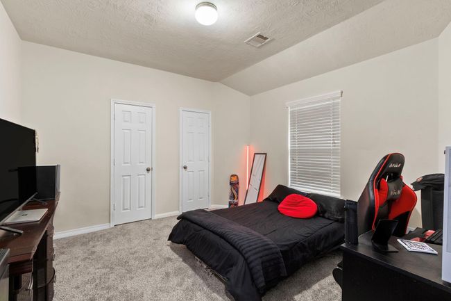 This room features a cozy space with neutral walls and carpeted flooring. It includes a bed, a gaming chair, and a desk setup, making it versatile for both relaxation and work. Two closets offer ample storage, and a window provides natural light. | Image 28