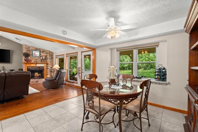 This photo showcases a cozy living and dining area with a warm, open layout. The space features a brick fireplace, hardwood and tile floors, and large windows offering a scenic view of the greenery outside. Both charm and functionality. | Image 7