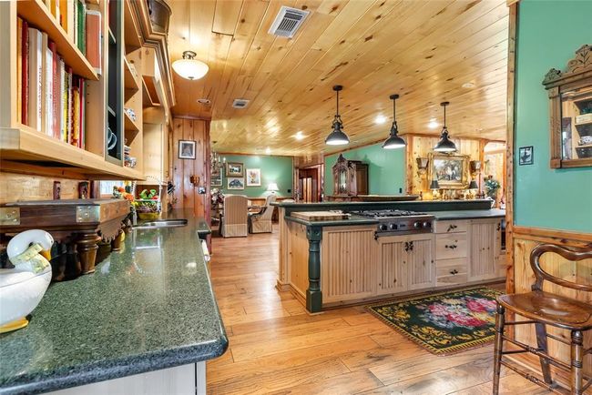 This cozy kitchen features warm wood paneling, a spacious island with a stove, and granite countertops. It opens into a charming dining area with elegant decor and ample natural light, creating a welcoming, rustic atmosphere. | Image 18