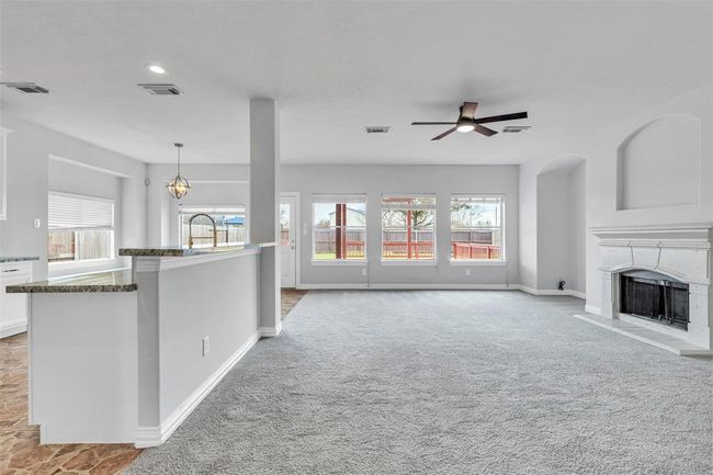 This spacious living area features a cozy carpeted floor, a modern ceiling fan, and a stylish fireplace. It opens up to a bright area with large windows offering a view of the backyard. The open layout includes a partial view of the kitchen with granite countertops. | Image 7