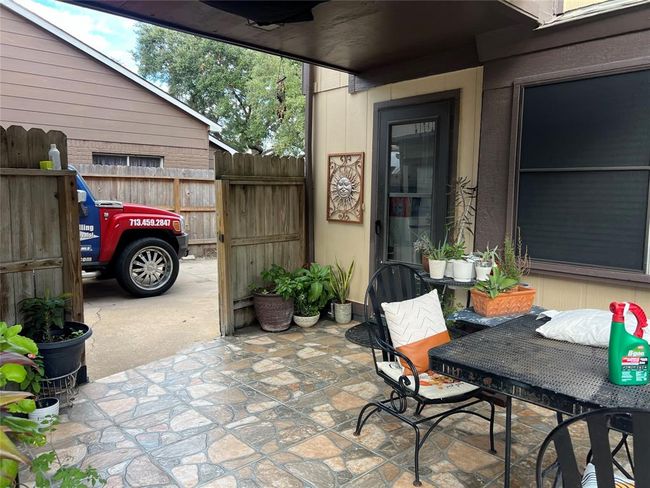 This photo shows a cozy outdoor patio area with tiled flooring and a small table with chairs. There are several potted plants adding greenery, and a wooden fence provides privacy. A door leads inside, and a parked vehicle is visible through an open gate. | Image 10