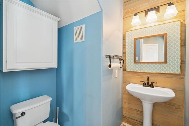 This bathroom features a cozy design with light blue walls, a pedestal sink, and a toilet. It includes a decorative mirror with a patterned frame, modern lighting, and a storage cabinet for convenience. | Image 26