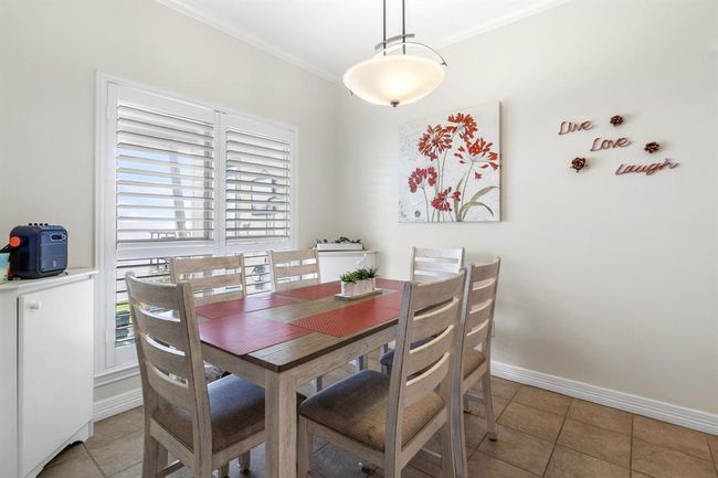 This dining area features a cozy, light-filled space with a modern table and six chairs. It has a bright window with plantation shutters, a stylish pendant light, and tasteful wall decor. | Image 10
