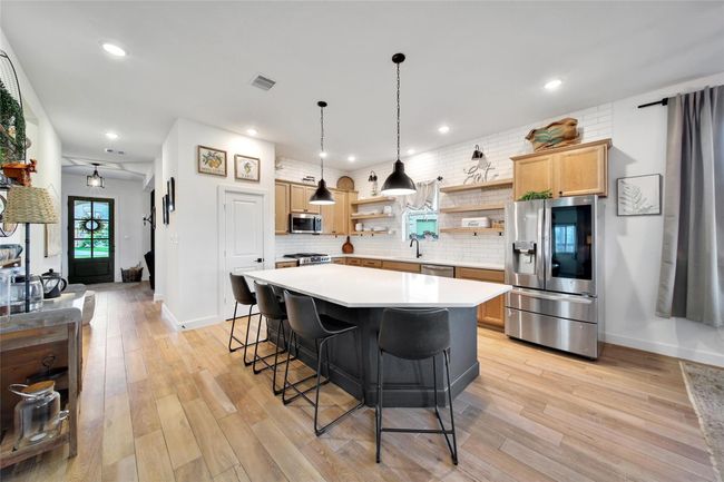 This kitchen features a spacious, modern design with a large island and seating for four. It has stainless steel appliances, light wood cabinetry, and open shelving. The room is bright and airy, with recessed lighting and stylish pendant lights. Wood flooring adds warmth, and a window over the sink provides natural light. | Image 17