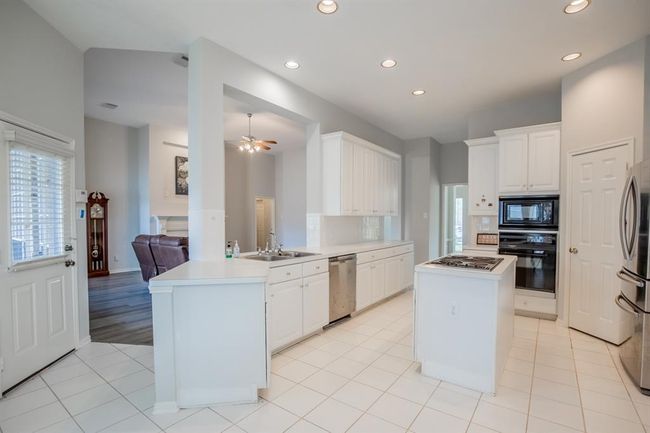 This spacious kitchen features clean white cabinetry, a central island with a cooktop. The open design connects seamlessly to a cozy living area with hardwood floors, creating a warm and inviting atmosphere. | Image 21