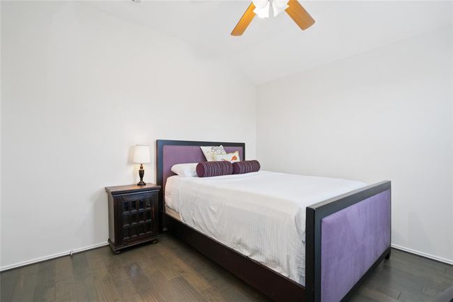 Cozy bedroom with a high ceiling, featuring a stylish bed with a purple headboard. Natural light and a ceiling fan enhance comfort, while a wooden nightstand offers practicality. | Image 41