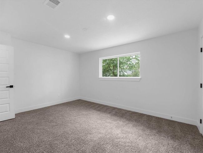 This room is spacious with neutral walls and plush carpet flooring. It features a large window allowing ample natural light and views of greenery. Recessed lighting and a modern door complete the look, offering a cozy and versatile space. | Image 29