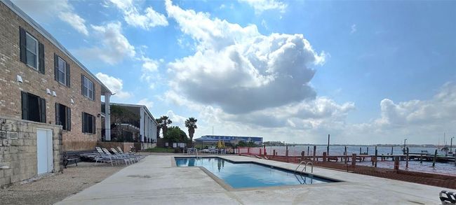 This photo showcases a waterfront property with a pool and a spacious deck area. The building features a brick exterior with multiple windows, and there are lounge chairs for relaxation. The view includes a large body of water and a partly cloudy sky, offering a serene and inviting atmosphere. | Image 30