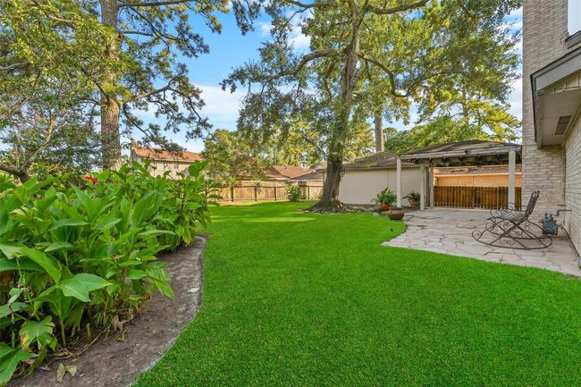 This photo showcases a spacious backyard with lush green grass, mature trees, and a charming patio area perfect for relaxation or entertaining. A wooden fence provides privacy, and there's a covered seating area for enjoying the outdoors. | Image 23