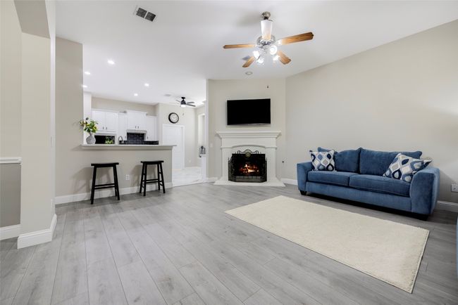 Living room featuring ceiling fan, a warm lit fireplace, light wood-type flooring, and view of the beautiful patio/yard. | Image 7