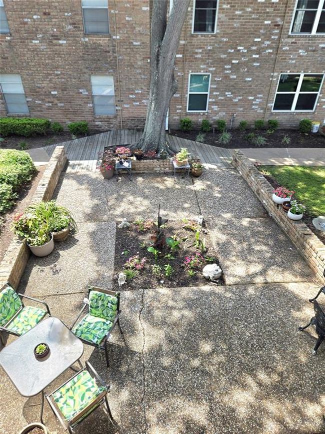 This charming outdoor space features a spacious stone patio with seating, surrounded by lush potted plants and flower beds. A mature tree provides natural shade, and the area is enclosed by a brick wall, offering privacy and a cozy atmosphere. Perfect for relaxing or entertaining. | Image 22