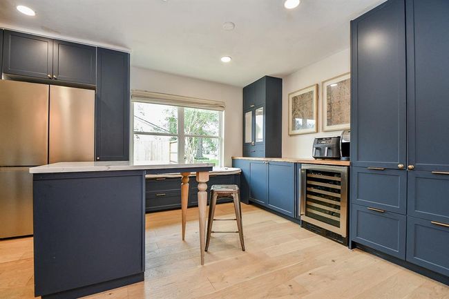 This kitchen features sleek, dark blue cabinetry with gold hardware, a modern stainless steel refrigerator, and a wine cooler. The wooden floors and large window provide a bright, inviting atmosphere, while a small island offers extra workspace. | Image 17