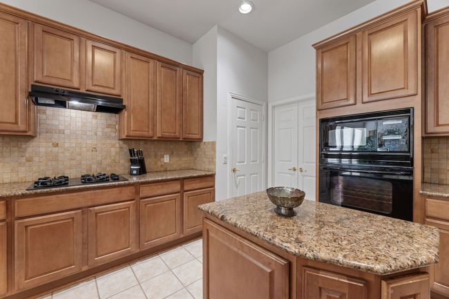 Kitchen with light tile patterned floors, backsplash, light stone counters, black appliances, and a kitchen island | Image 16