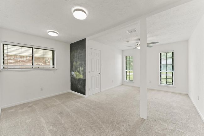 This room features a clean, open layout with neutral carpeting and white walls. It has ample natural light from multiple windows, recessed lighting, and a ceiling fan. A partition provides division, and there's a small chalkboard wall for creative use. | Image 22
