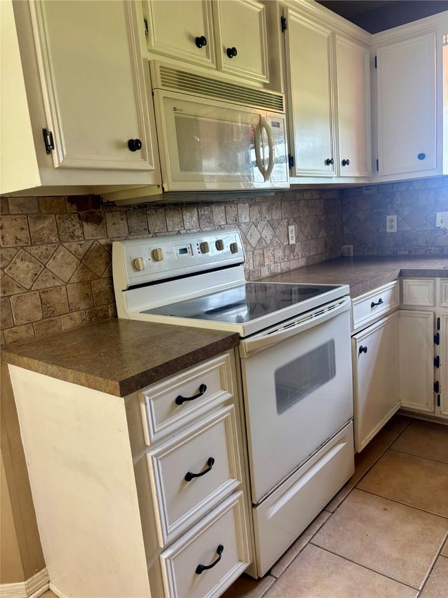 Kitchen with white appliances, dark countertops, backsplash, and light tile patterned floors | Image 9