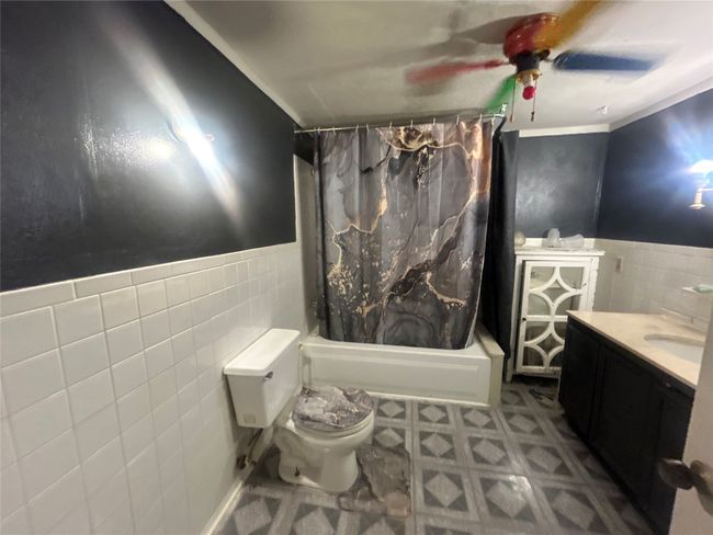 Bathroom with tile walls, shower / bath combo with shower curtain, a ceiling fan, vanity, and a wainscoted wall | Image 8