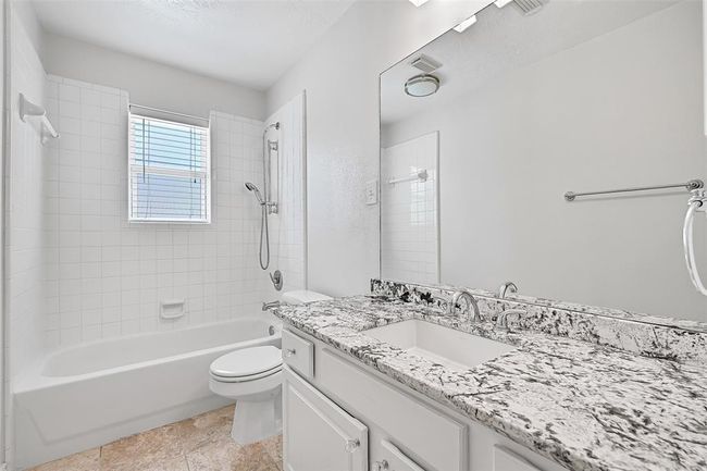 The updated hall bathroom features a sleek granite countertop with an undermount sink, chrome fixtures, white cabinetry, tile floors, and a tub/shower combo with a classic white tile surround and a handheld showerhead. | Image 20