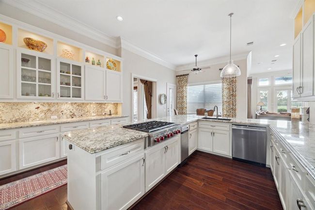 This spacious kitchen features elegant granite countertops, white cabinetry with glass accents, and modern stainless steel appliances. It has a central island with a gas cooktop, ample storage, and pendant lighting. The open layout connects seamlessly to a bright dining area with large windows. | Image 13
