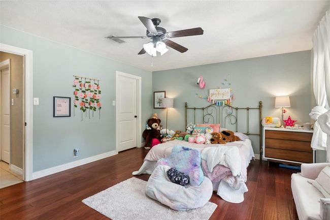 This cozy bedroom features soft blue-green walls and warm hardwood floors, creating a calming atmosphere. It includes a ceiling fan, ample natural light from a large window, and tasteful decor. Ideal for a child's room, it has plenty of space for toys and personal touches. | Image 35