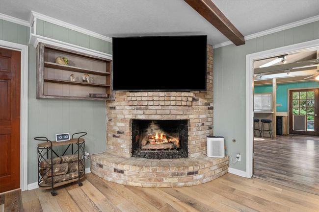 Cozy living area featuring a rustic brick fireplace with an area to have your TV mounted above. The space includes beautiful flooring, a built-in shelf, and opens to a game/ flex room with large windows and wood details- Perfect for relaxation and entertaining! | Image 16