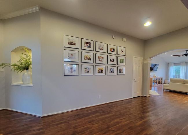This photo showcases a spacious living area with elegant wood flooring and neutral walls. It features a gallery wall with framed photos and an arched entryway leading to a well-lit dining and living space. A decorative niche with a vase and greenery adds charm to the room. | Image 15