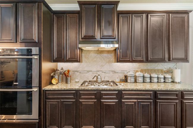 This kitchen features rich, dark wood cabinetry and granite countertops, offering ample storage and workspace. It includes a built-in double oven and a gas cooktop with a stylish range hood. The backsplash adds a subtle decorative touch, complementing the overall elegant design. | Image 10