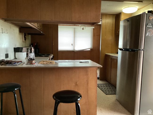 Kitchen with freestanding refrigerator, a peninsula, light countertops, a breakfast bar area, and brown cabinetry | Image 9