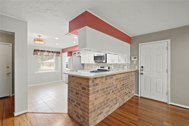 This photo showcases a bright, open kitchen with modern appliances and white cabinetry. The space features a brick accent bar with tile flooring in the kitchen and recessed lightening. Door pictured leads to the 2 car garage. | Image 8