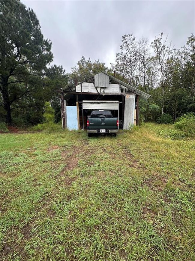 This photo shows a rustic, metal-roofed garage or shed with a vehicle parked inside, surrounded by a grassy area and trees, offering a secluded and natural setting. | Image 12