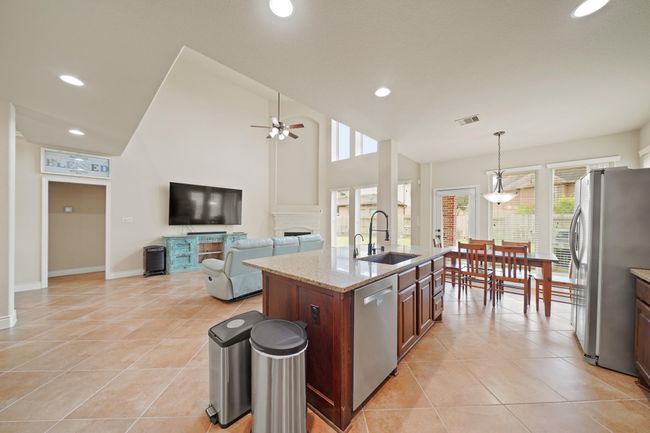 This open-concept living area features a high ceiling with ample natural light. It includes a spacious kitchen with a large island, modern appliances, and a dining area. The living room has a ceiling fan and fireplace, creating a comfortable and inviting space. | Image 17