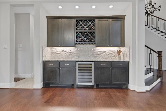 A stylish home dry bar area with dark wood cabinets, a built-in wine rack, and a sleek wine cooler. The backsplash features mosaic stone tiles, and recessed lighting enhances the elegant design. Adjacent to stairs, it offers a modern, inviting space ideal for entertaining. | Image 14