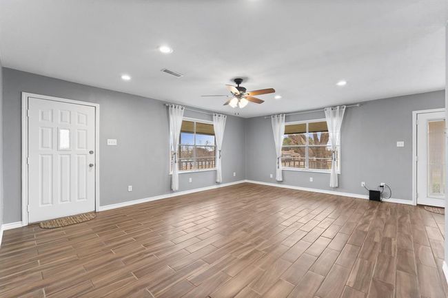 This spacious living room features wood-look tile flooring, neutral gray walls, and ample natural light from large windows with white curtains. It includes a ceiling fan and recessed lighting, creating a bright and airy atmosphere. | Image 10