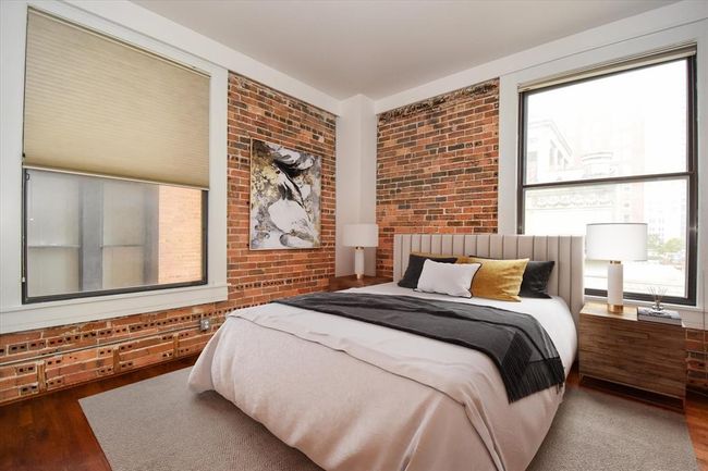 This inviting bedroom features classic exposed brick, large windows for natural light, and a modern design with warm wood floors, perfect for downtown living. | Image 7