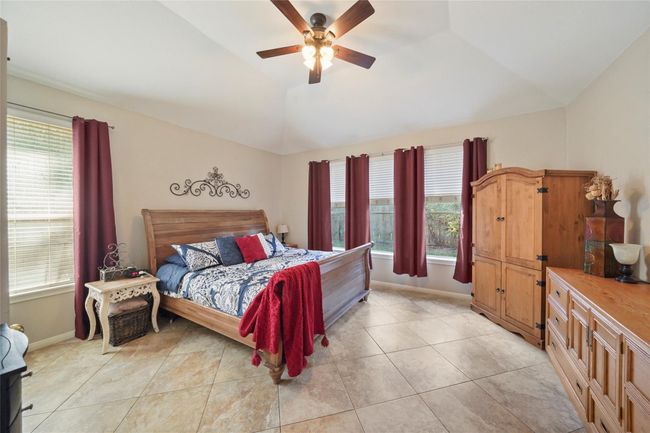 This spacious bedroom features a vaulted ceiling with a ceiling fan, large windows with burgundy curtains providing natural light. The room is accented with tile flooring. | Image 18