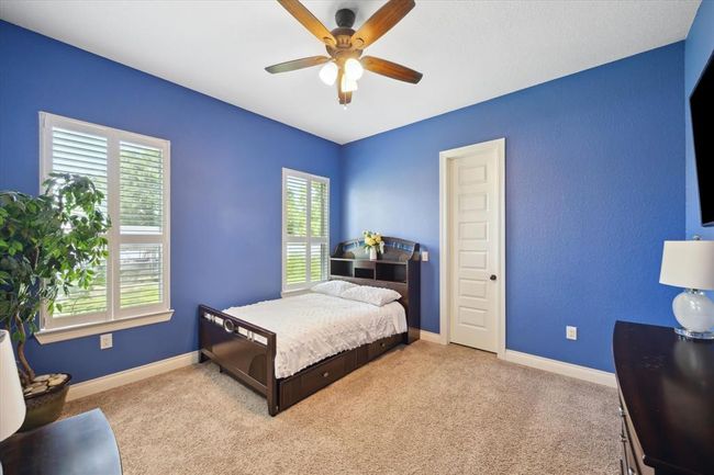 This bedroom features vibrant blue walls, a cozy carpeted floor, and ample natural light from two large windows. It includes a ceiling fan, a bed with a dark wood frame, and a door leading to another room or closet, creating a comfortable and inviting space. | Image 11