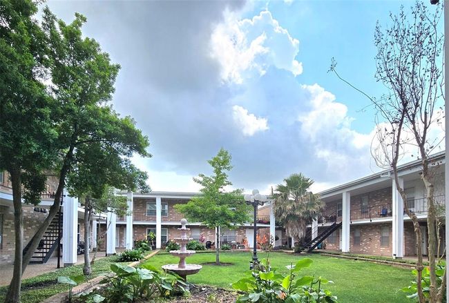 This photo showcases a charming courtyard in a residential complex, featuring well-maintained greenery, a central fountain, and surrounding two-story buildings with exterior staircases, creating a welcoming and serene atmosphere. | Image 28
