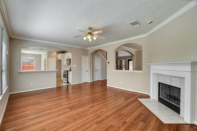 This spacious living area features warm wood flooring, a cozy fireplace, and abundant natural light. The open layout flows into the kitchen, separated by stylish archways, creating an inviting and functional space for family living. | Image 15