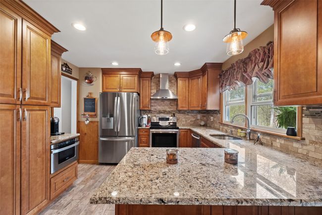 Kitchen featuring a sink, stainless steel appliances, brown cabinetry, wall chimney exhaust hood, and light stone counters | Image 10