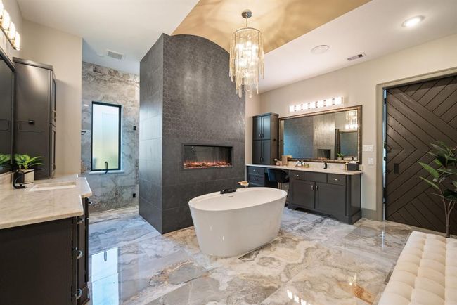 This luxurious bathroom features a freestanding tub, a modern fireplace, and elegant marble flooring. It includes dual vanities, a large mirror, and a stylish chandelier, creating a spa-like atmosphere. | Image 30