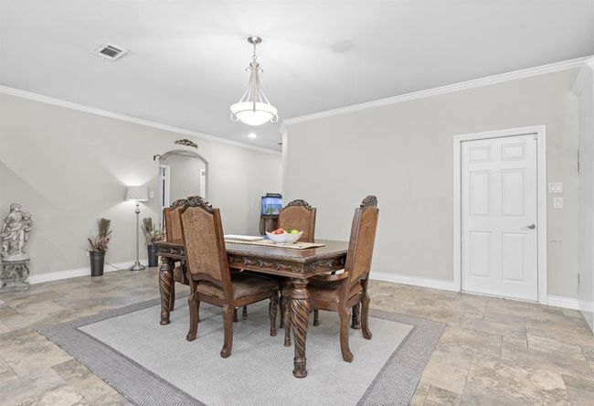 This is a spacious dining room featuring a large, ornate wooden table with chairs, tiled flooring, soft neutral wall colors, ample lighting, and a classic white door. The room is accessorized with a statue and a floor lamp, creating an elegant atmosphere. | Image 20