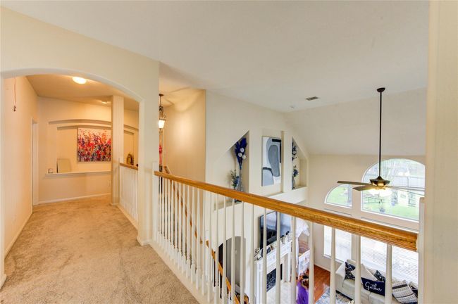 This photo showcases a bright upper-level hallway with carpeted floors and an open view of the living area below. The space features arched accents, a railing with wooden trim, and large windows that let in natural light. There's also decorative wall art and a ceiling fan visible in the adjacent room. | Image 28