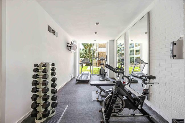 The gym features modern equipment, including dumbbells, a stationary bike, treadmills, and large mirrors, providing a bright and functional workout space with views of the outdoors. | Image 22