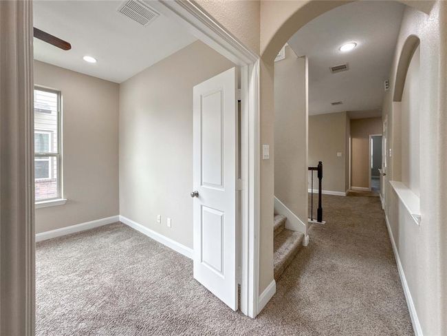 This photo showcases a cozy carpeted hallway with an adjacent room featuring natural light from a window. The hallway leads to a staircase and additional rooms, creating a warm and inviting environment suitable for a comfortable home. | Image 28