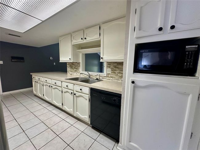 This kitchen features white cabinetry with a contrasting dark backsplash and black appliances. It includes ample counter space, a double sink, and a built-in microwave. The tile flooring and overhead lighting create a bright and functional cooking area. | Image 7