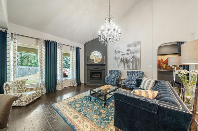 This spacious living room features high vaulted ceilings, elegant dark wood flooring, and a cozy fireplace. Large windows with stylish curtains provide plenty of natural light. The room is tastefully decorated with a chandelier, offering a warm and inviting atmosphere. | Image 6