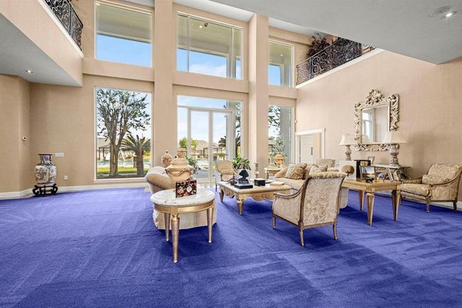 This is a spacious living room with high ceilings, large windows providing ample natural light, and a view of the outdoors. It features a bold purple carpet and classic-style furniture, with a second-story indoor balcony overlooking the space. | Image 8