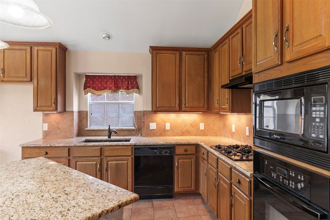 Kitchen with black appliances, a sink, brown cabinetry, under cabinet range hood, and light stone countertops | Image 27