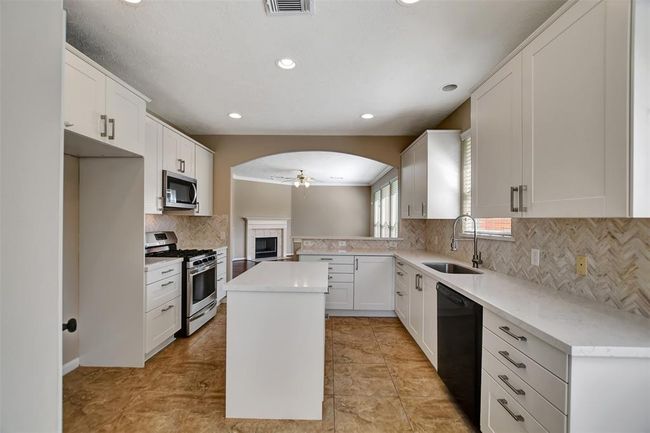 This bright, modern kitchen features white cabinetry, a central island, and stainless steel appliances. The space opens to a living area with a cozy fireplace, and large windows provide ample natural light. | Image 22