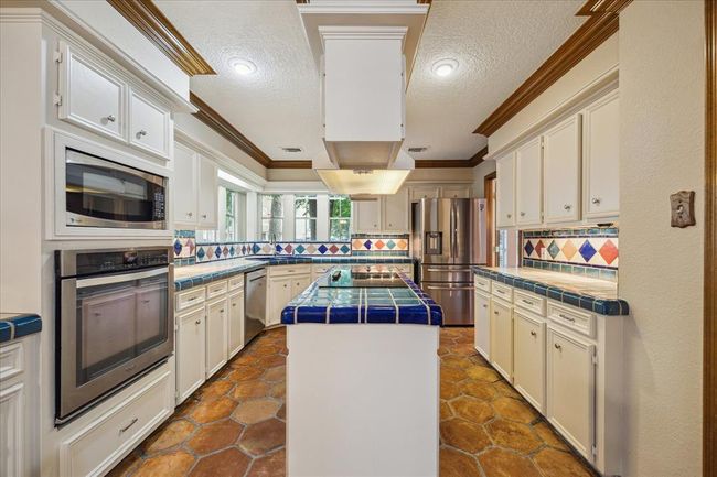 This kitchen features classic white cabinetry, vibrant tile countertops, and a multi-colored tile backsplash. It includes modern stainless steel appliances, a central island, and saltillo tile flooring, creating a warm and inviting cooking space. | Image 10